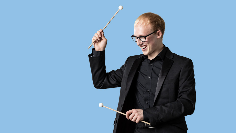 Student in a black suit holding percussion beaters, against a light blue background